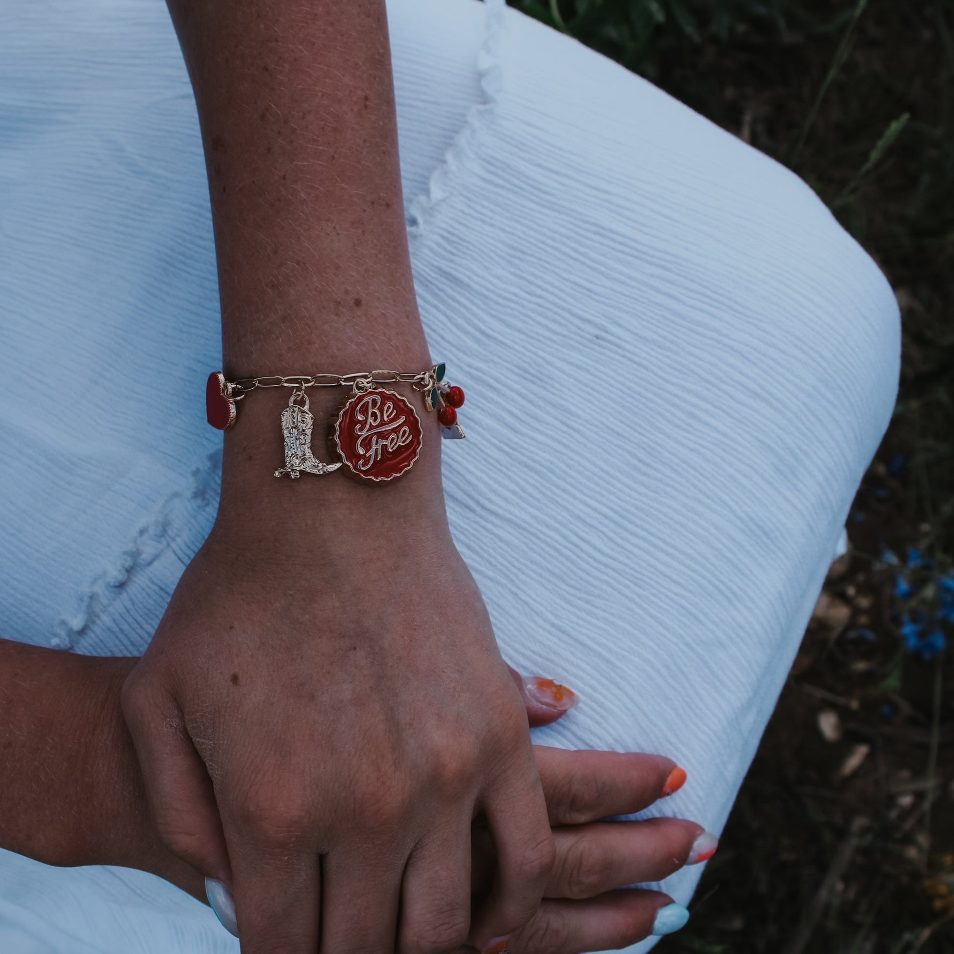 Person wearing a bracelet with a red charm on a white surface. Cowgirl bracelet. boots. heart. cherry.