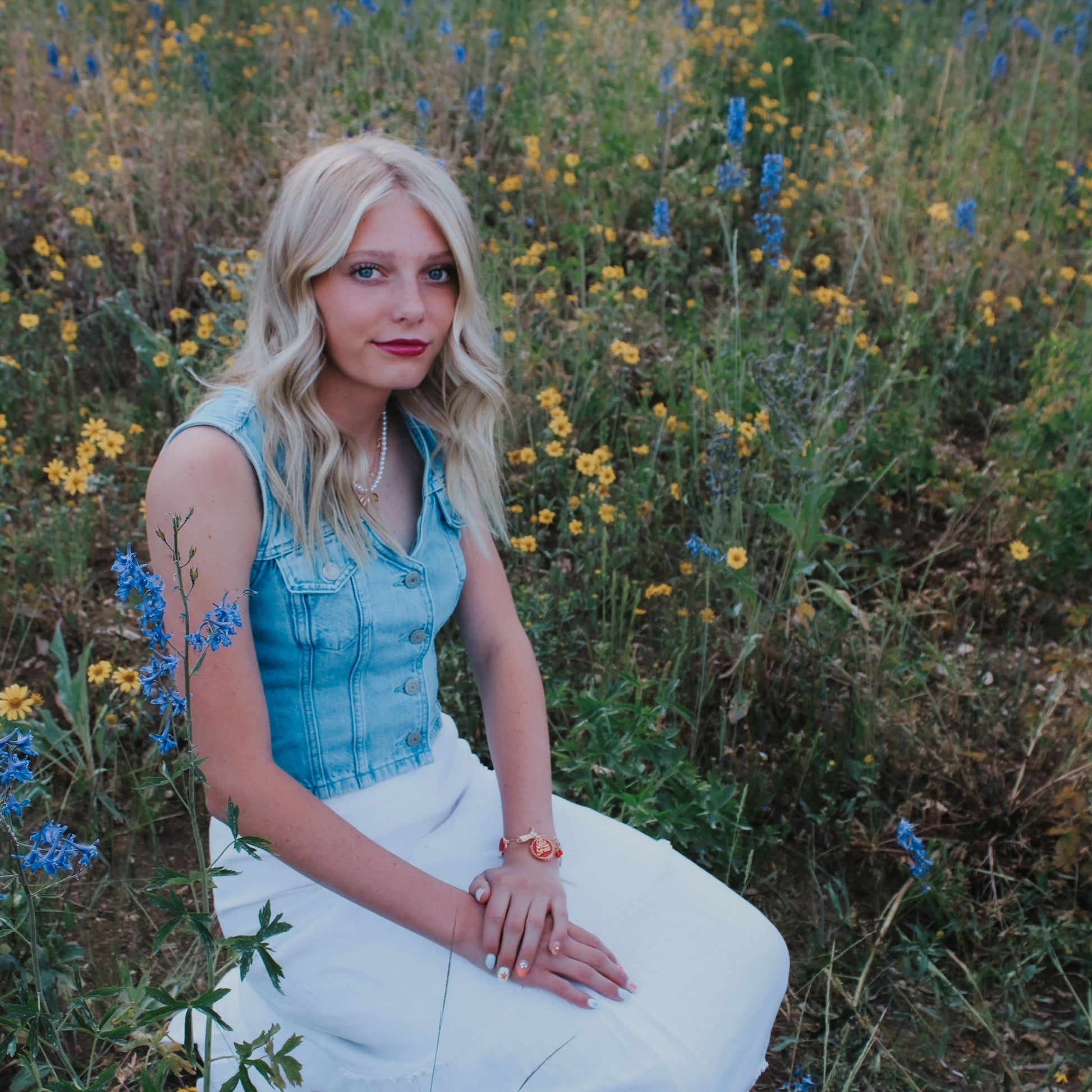 Woman sitting in a field of wildflowers wearing a blue top and white pants. Charm bracelet. be free. red. cherries. cowgirls boots western 